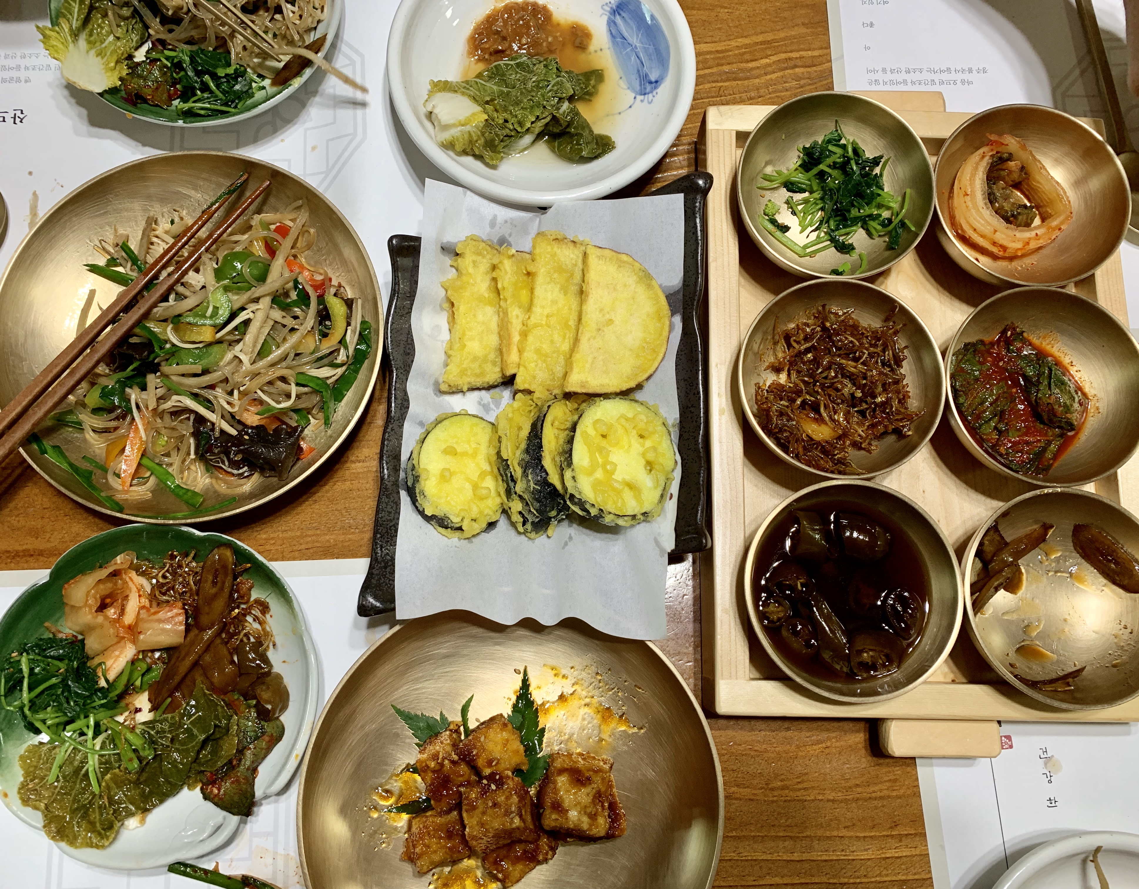 Three girls sitting on the floor of a wood-paneled room, in front of rows of red velvet seats. The girl on the left wears a white T-shirt, a green sweatshirt and glasses and holds a pen and a piece of paper. The girls on the right wear a white T-shirt and a blue T-shirt and pink shorts and gesture toward the piece of paper.